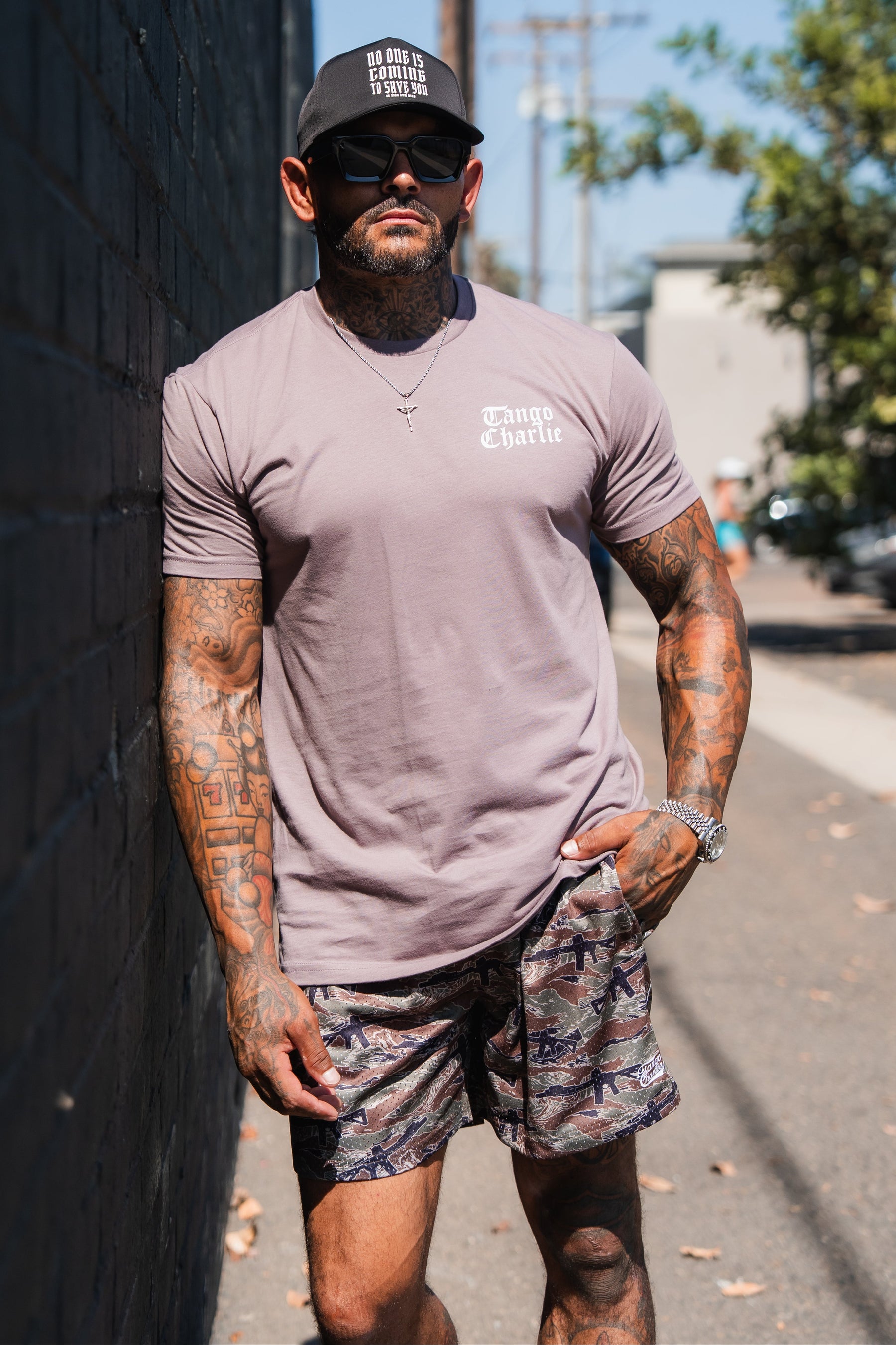 A tattooed man wearing a mauve Lift Heavy. Praise Hard. Shiitake Tee, camouflage shorts, sunglasses, and a baseball cap leans against a brick wall.