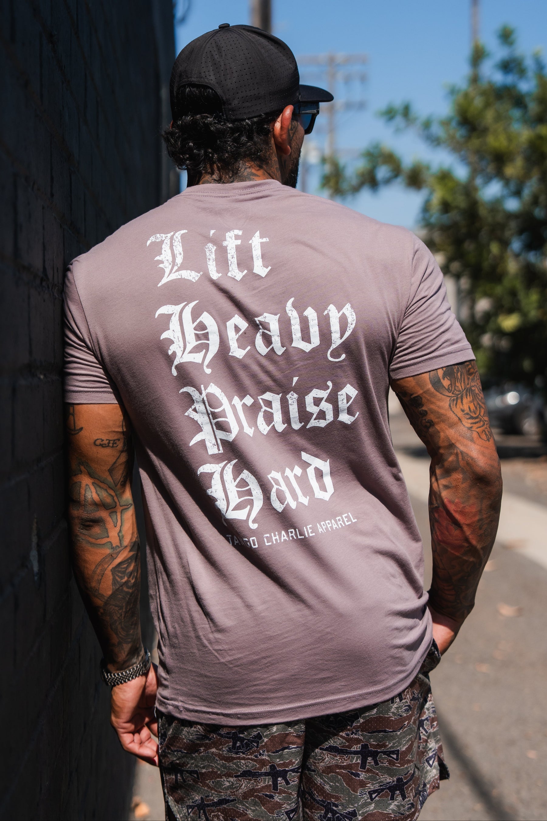 A man wearing a mauve Lift Heavy. Praise Hard. Shiitake Tee with white gothic-style lettering on the back.