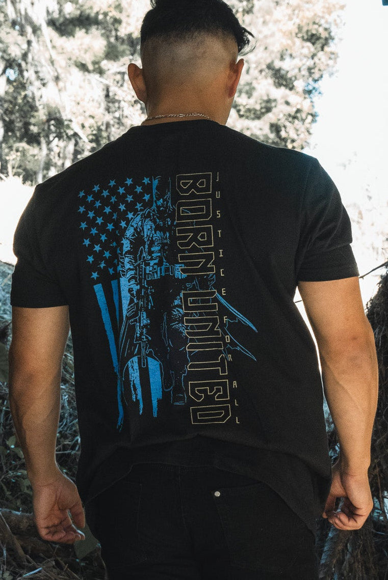 A person with short hair wearing Born United Private Labels Justice For All original printed tee strolls under a structure along a natural path, surrounded by foliage. With their back to the camera, they explore the outdoors like one of Gothams masked heroes in an urban jungle.