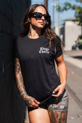 A person wearing a Lift Heavy. Praise Hard. Black Tee leans against a brick wall, the black t-shirt featuring Praise Hard printed in white gothic-style text on the chest.