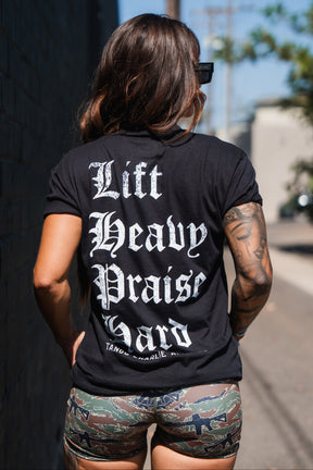 A woman seen from behind wearing a black Tango Charlie Apparel Lift Heavy. Praise Hard. Black Tee with white gothic lettering inspired by Isaiah 41:10.