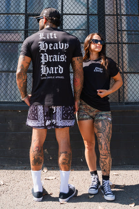 A man and a woman in black t-shirts stand before a chain-link fence; the man wears a Lift Heavy. Praise Hard. Black Tee by Tango Charlie Apparel, with the text visible on the back.