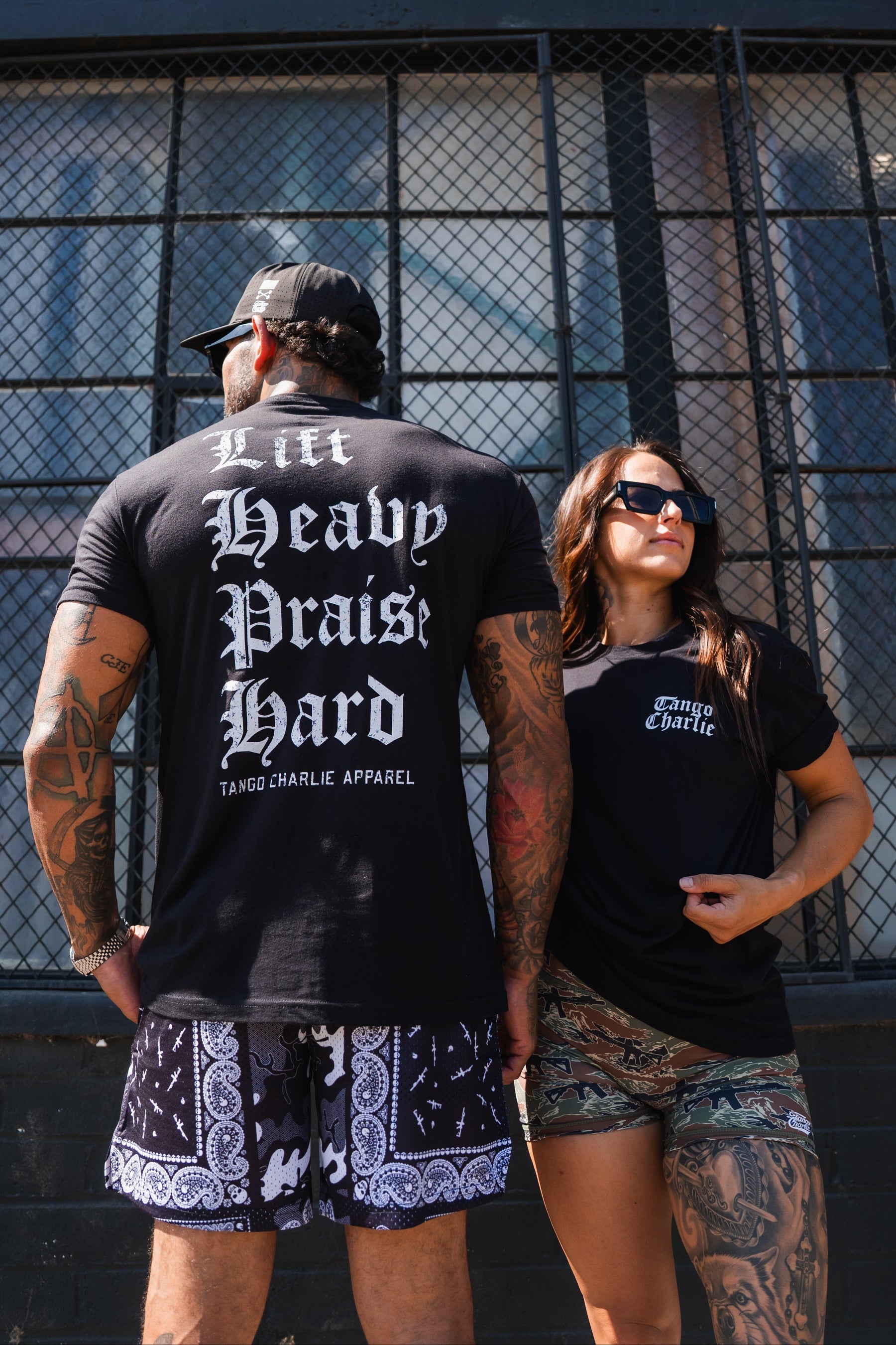 A man and woman standing in front of a chain-link fence, wearing black Lift Heavy. Praise Hard. Black Tee shirts.
