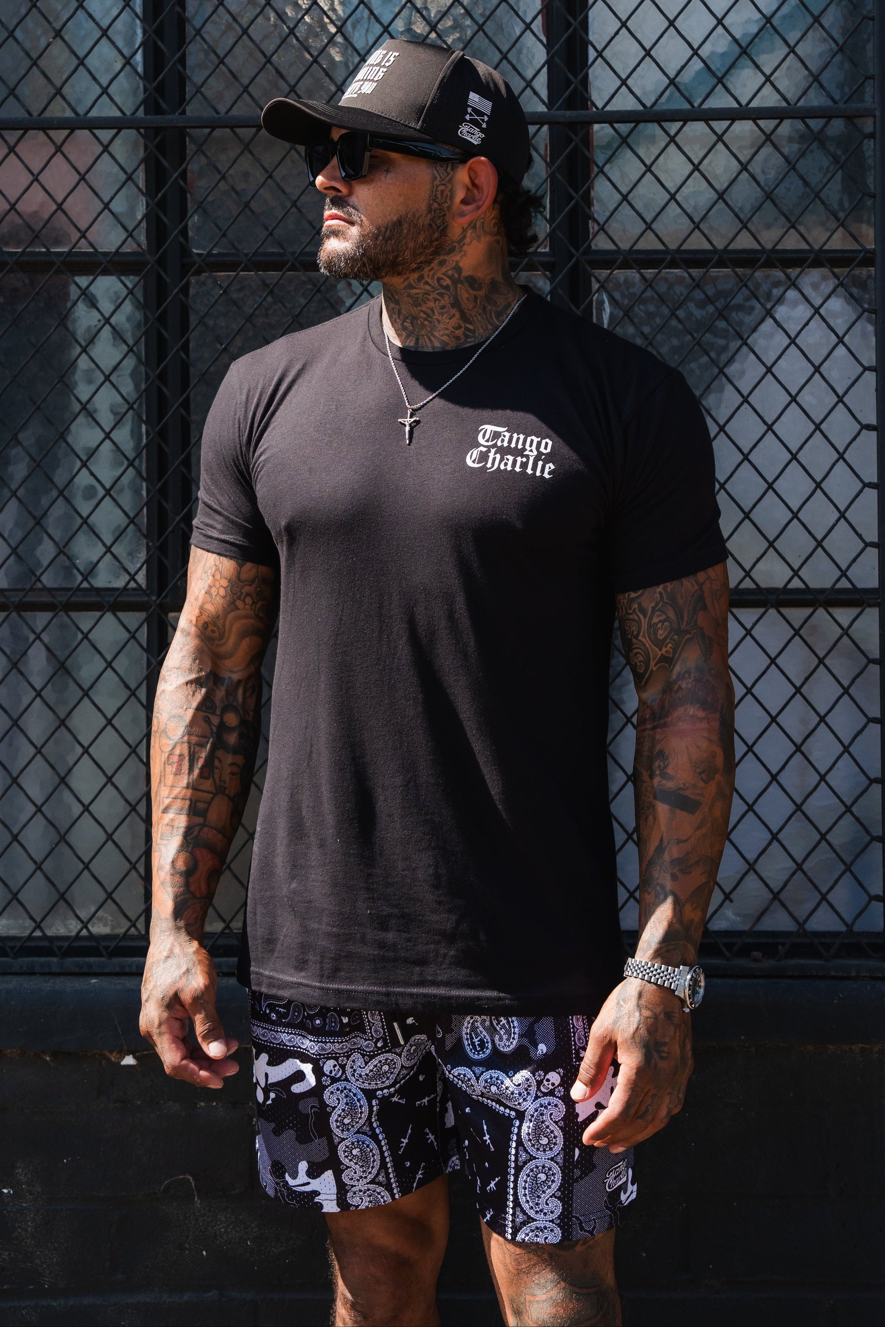A tattooed man wearing a black baseball cap, sunglasses, patterned shorts, and a Lift Heavy. Praise Hard. Black Tee by Tango Charlie Apparel.