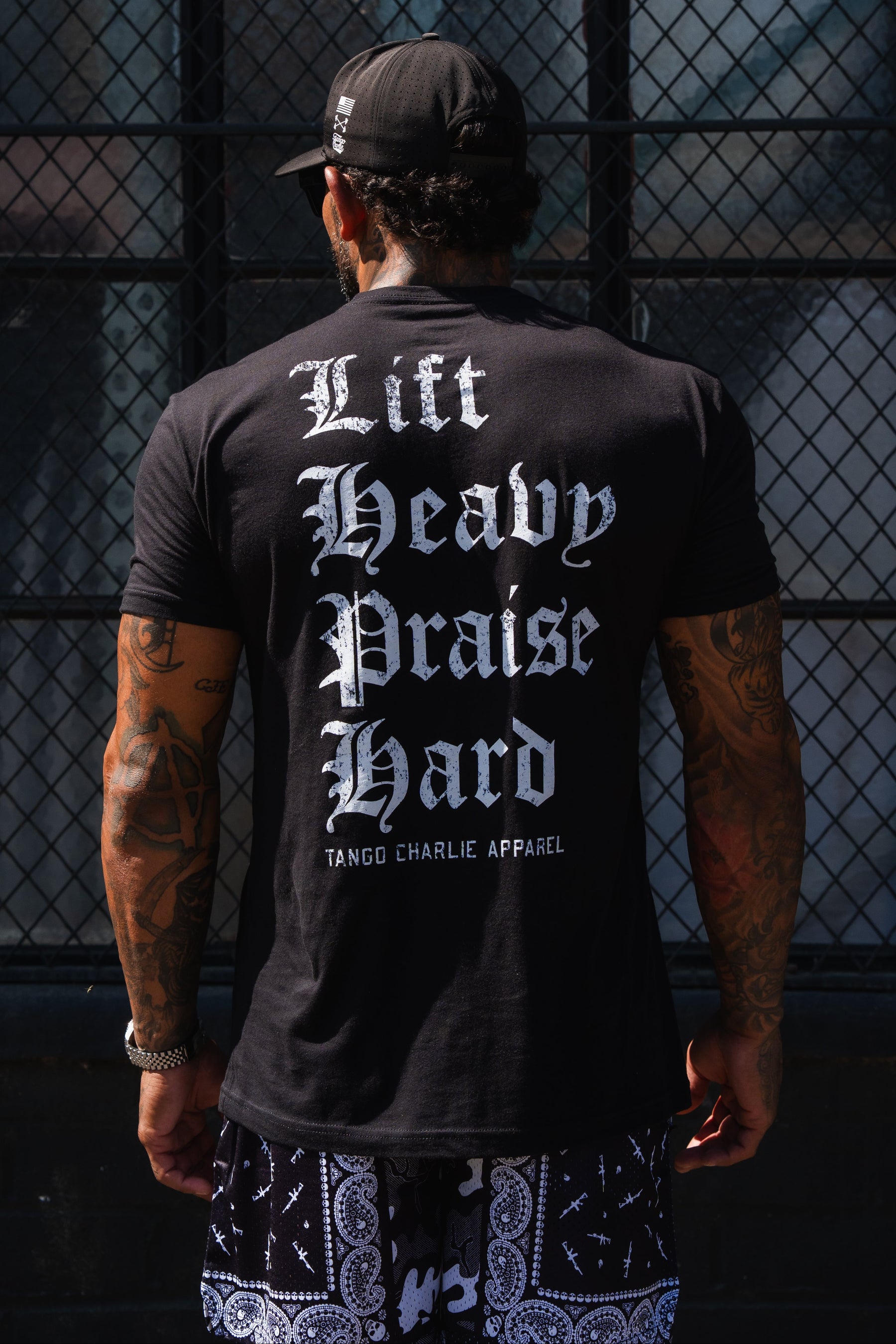 Back view of a person wearing a black Tango Charlie Apparel Lift Heavy. Praise Hard. Black Tee with white gothic lettering inspired by Isaiah 41:10.