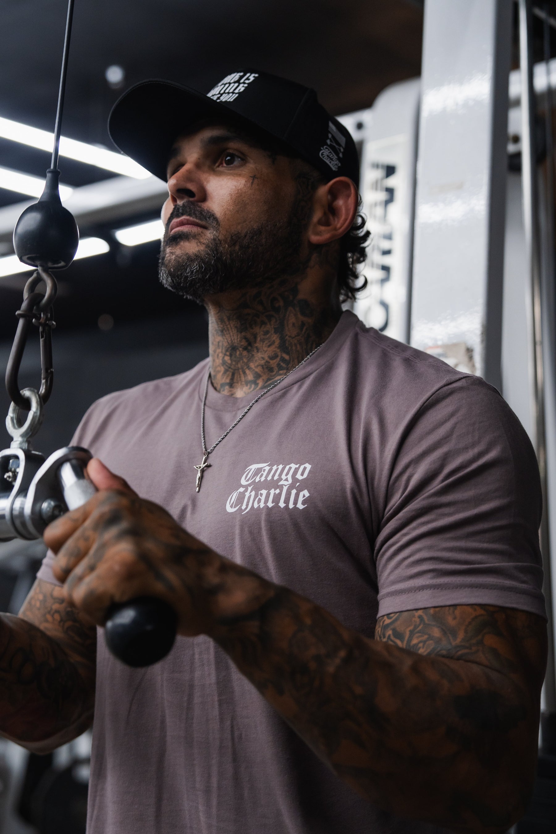 A tattooed man in a mauve Lift Heavy. Praise Hard. Shiitake Tee and baseball cap performs a cable pulldown in the gym.