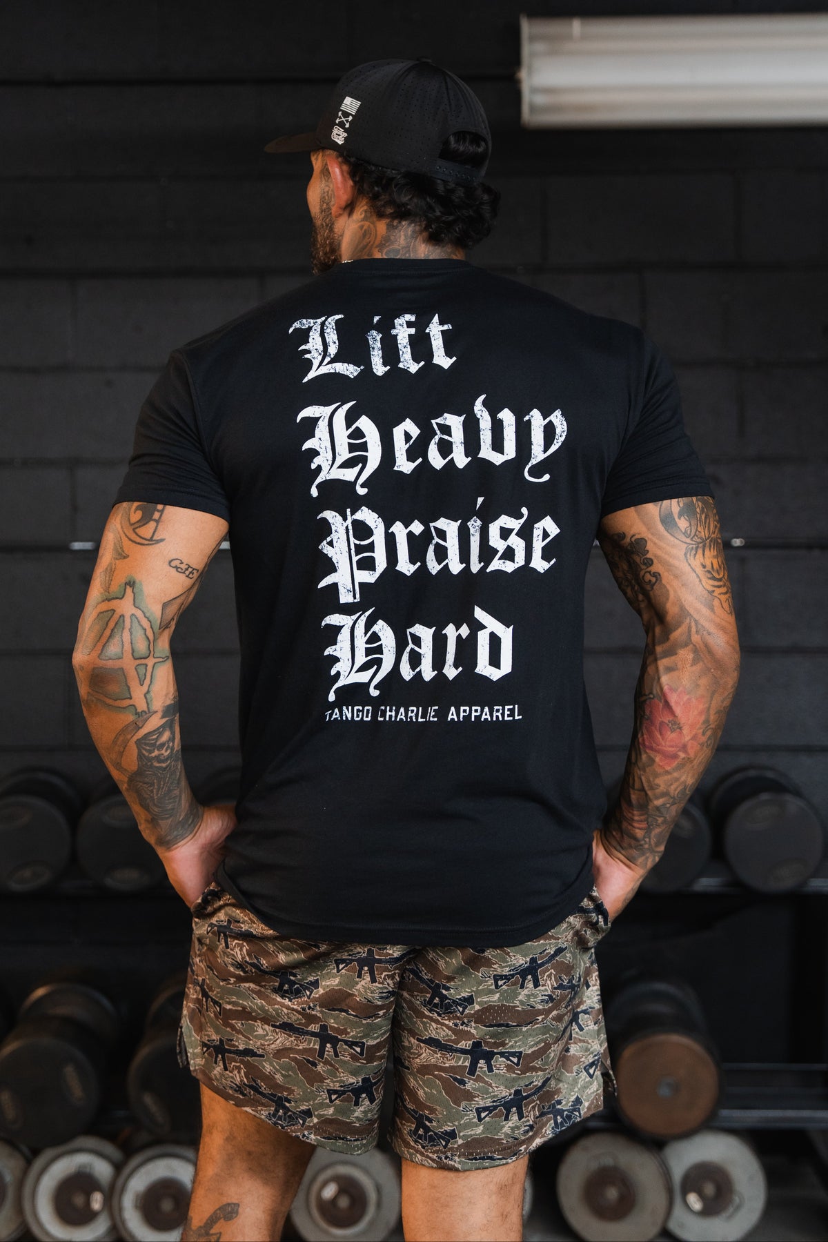 A person seen from behind wearing a black Lift Heavy. Praise Hard. Black Tee with the words printed in white gothic-style lettering.