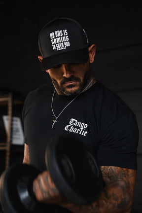 A man with tattooed arms lifts heavy while curling a dumbbell, wearing a black cap that reads No one is coming to save you and a Lift Heavy. Praise Hard. Black Tee from Tango Charlie Apparel.