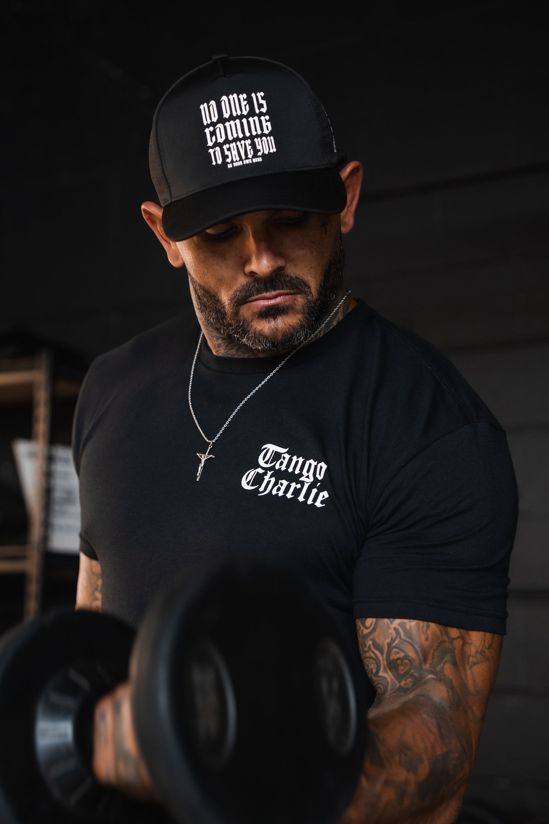 A man wearing a Lift Heavy. Praise Hard. Black Tee and a black baseball cap with the text No one is coming to save you lifts a heavy dumbbell.