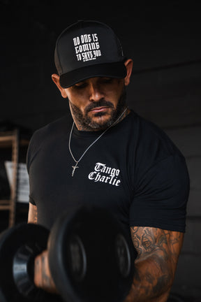 A man wearing a Lift Heavy. Praise Hard. Black Tee and a black baseball cap with the text No one is coming to save you lifts a heavy dumbbell.