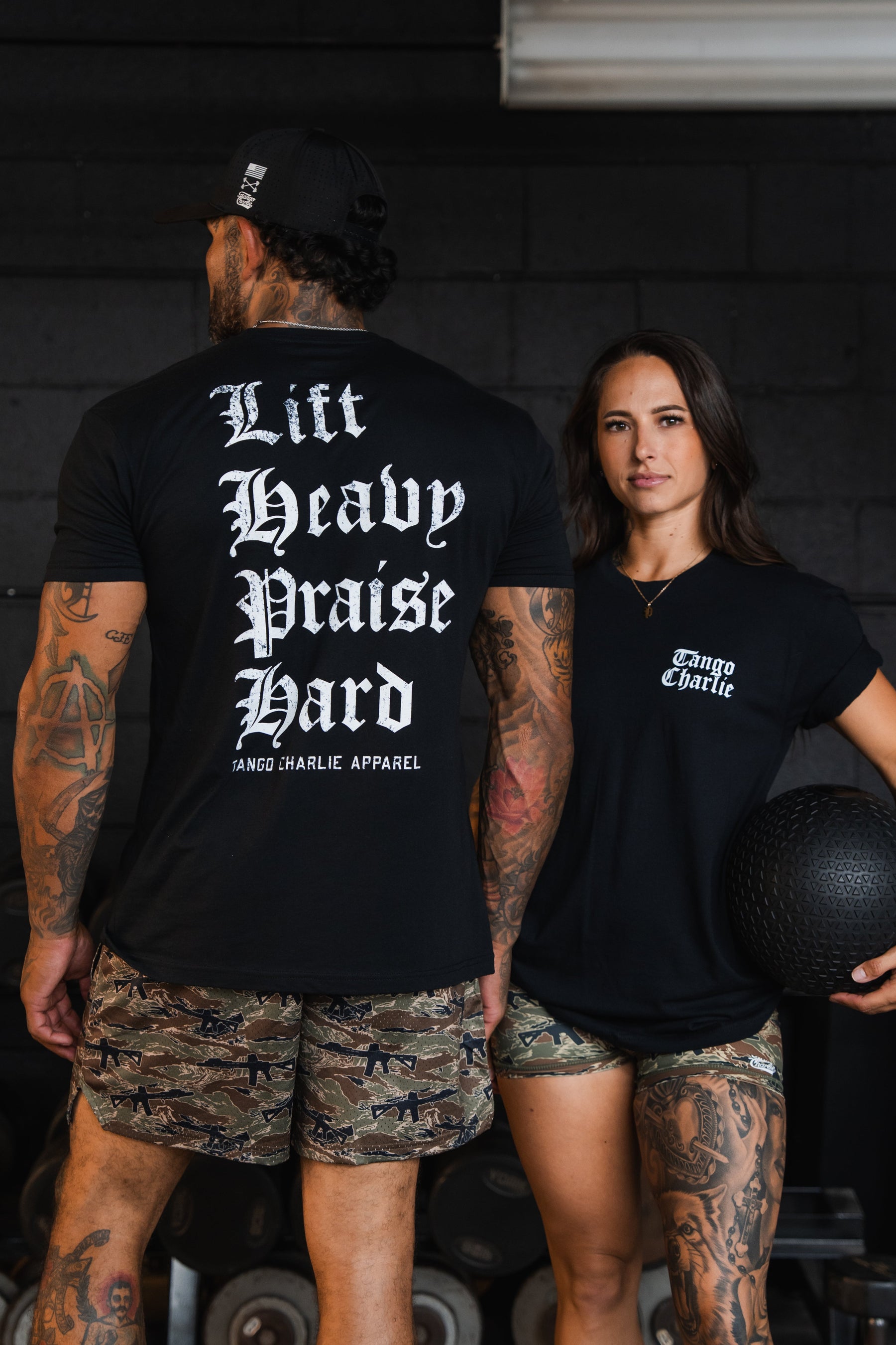 A man and woman stand in a gym wearing black Tango Charlie Apparel Lift Heavy. Praise Hard. Black Tee shirts; the back of the man’s shirt displays the message to lift heavy and praise hard.
