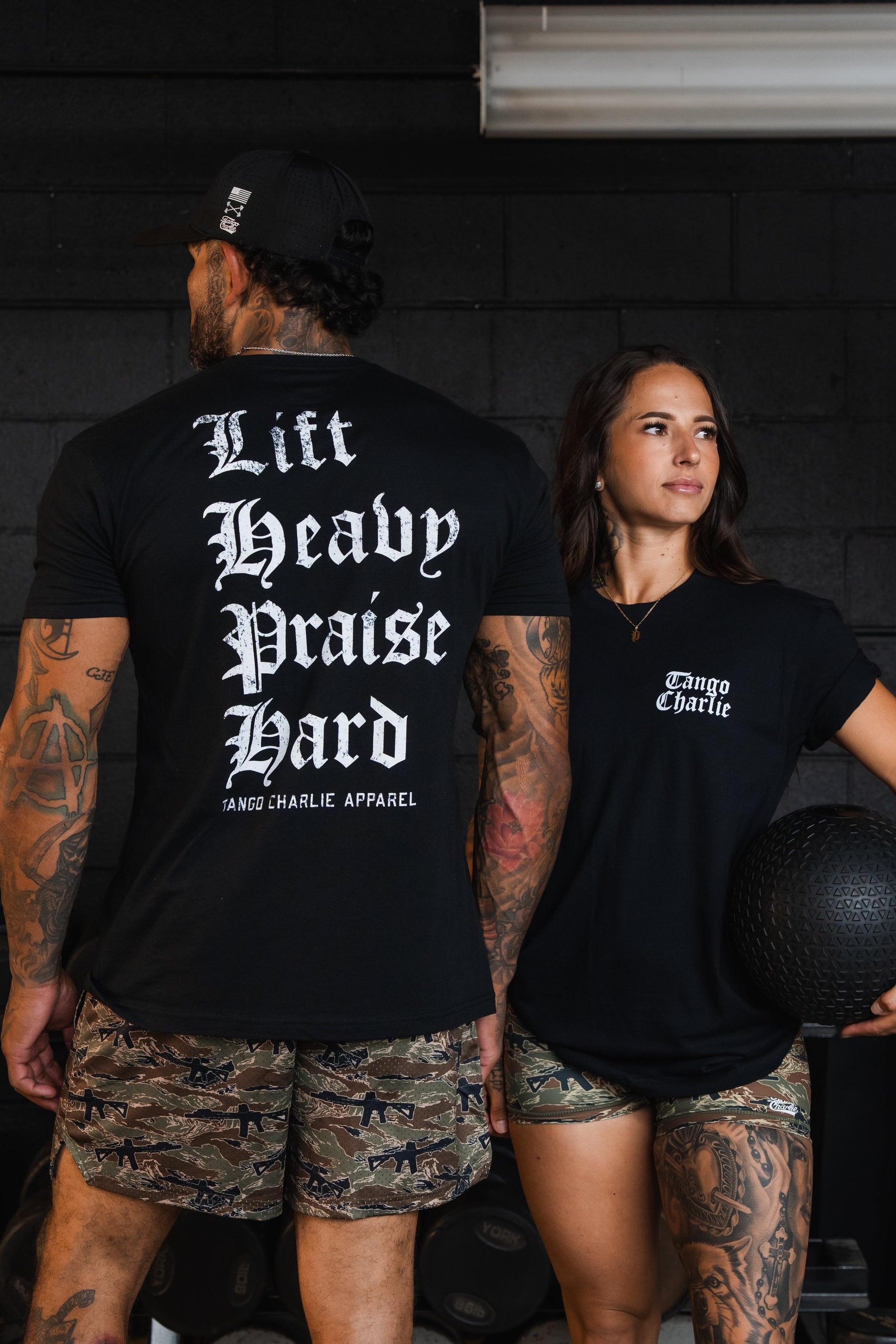 A man and woman in a gym wearing black Tango Charlie Apparel Lift Heavy. Praise Hard. Black Tees with the text on the back and chest.