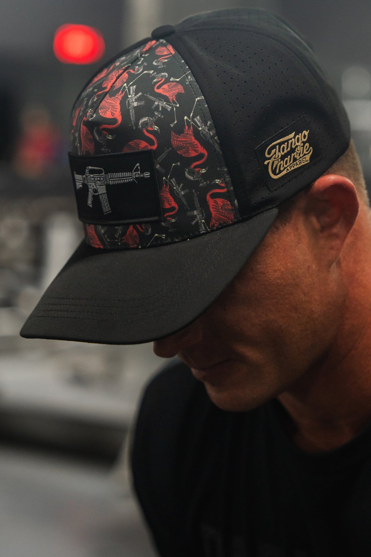Wearing a black Tactimingo Hydro snapback by Tango Charlie Apparel, the individual shows off red flamingos and a rifle patch on this sweat-resistant cap with a blurred background.