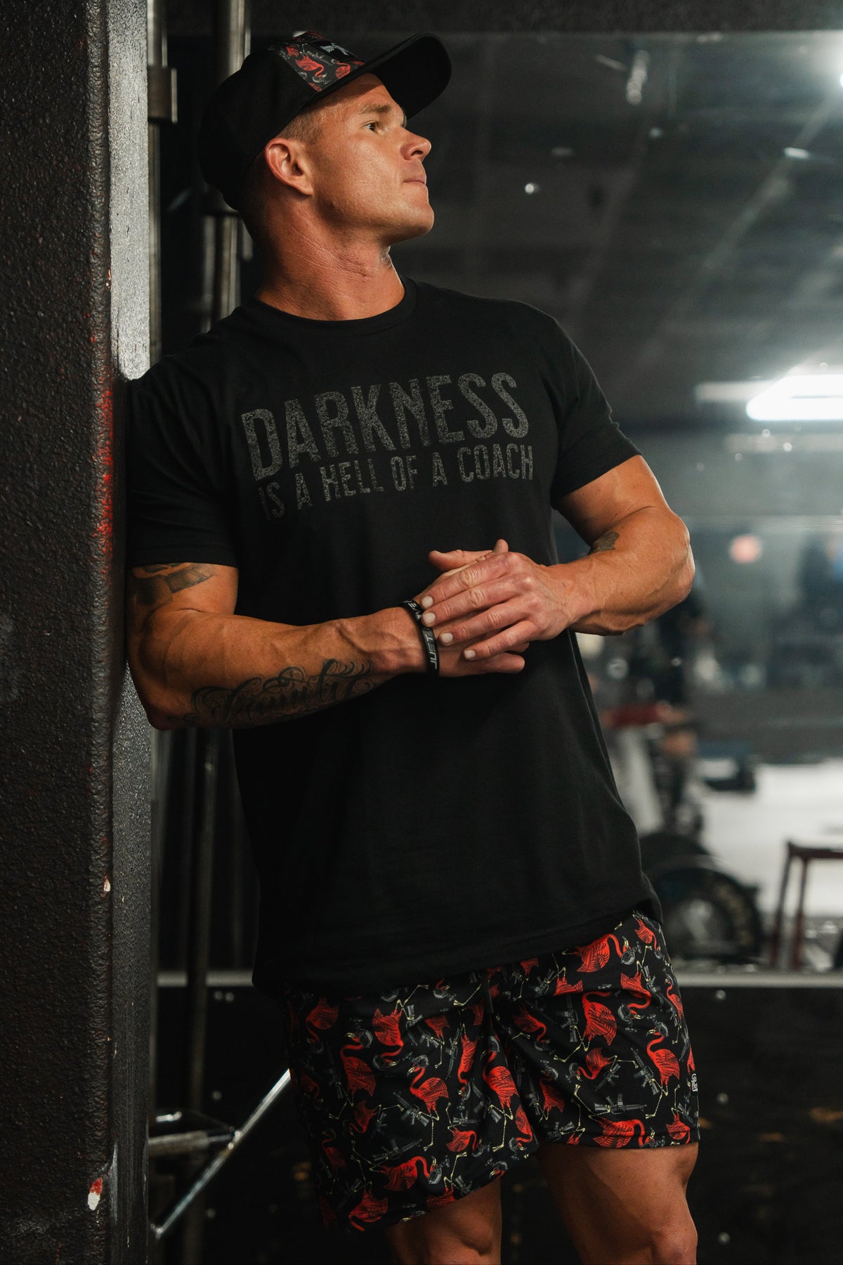 In a dimly lit gym, someone leans against the wall, wearing a black cap, dark shirt with DARKNESS IS A HELL OF A COACH, and Tango Charlie Apparels Mens Performance Training Shorts | 5.5 - Tactimingo. Tattoos on their left arm enhance their intense and focused demeanor.