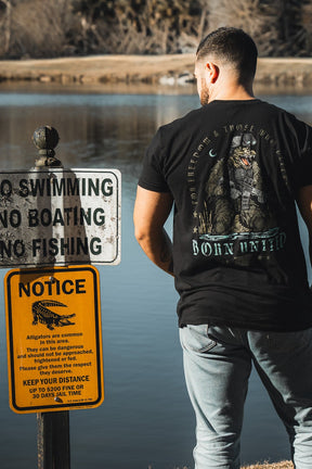 A man in jeans and the Born United Black Box - Tactigator tee by Born United Private Label stands by a lake near signs that prohibit swimming, boating, and fishing, and warn of dangerous alligators.