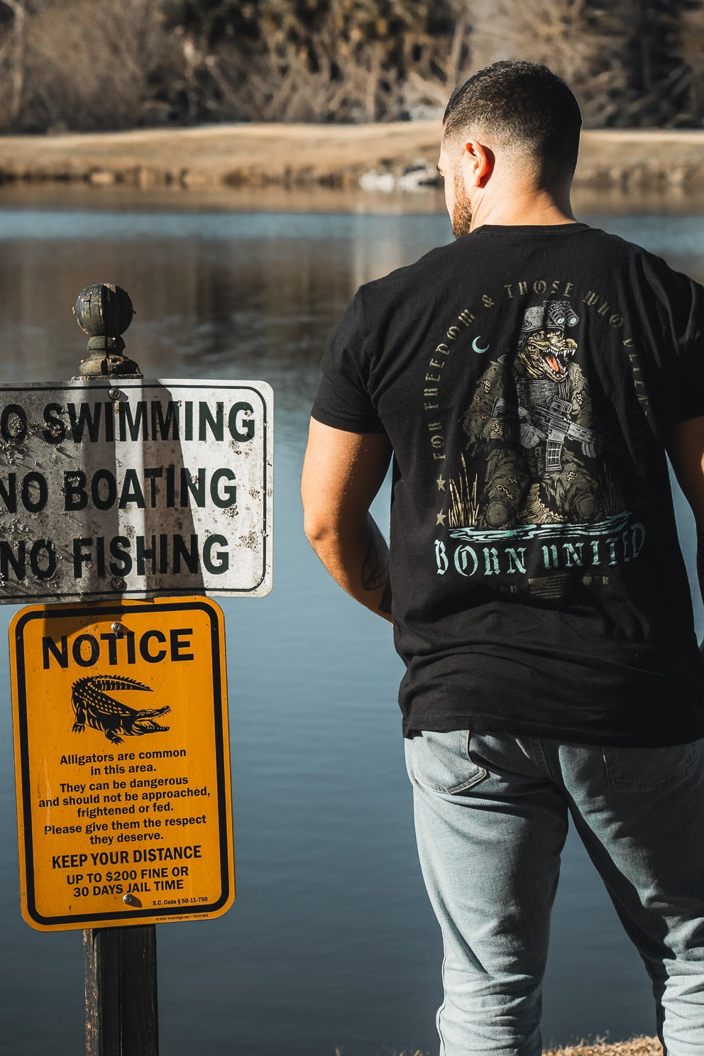 A man in jeans and the Born United Black Box - Tactigator tee by Born United Private Label stands by a lake near signs that prohibit swimming, boating, and fishing, and warn of dangerous alligators.
