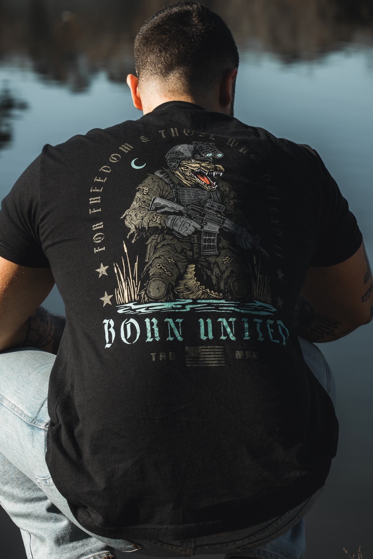 A man crouches by water, seen from behind, wearing the Born United Private Label Black Box - Tactigator tee featuring a bear in tactical gear and the phrases "Born United" and "For Freedom & Those Who Defend" on the back.
