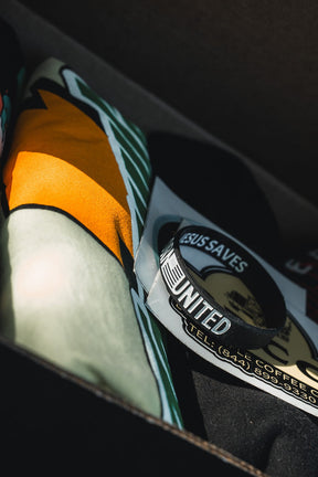 A close-up of the Born United Black Box - Tactigator by Born United Private Label shows a colorful folded fabric, a JESUS SAVES sticker, and a black-and-white UNITED bracelet, all partially lit by sunlight.