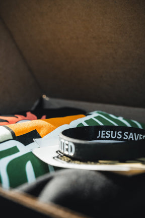 A close-up of a black JESUS SAVES wristband on colorful fabric and cards inside the Born United Private Label’s Tactigator Black Box, with sunlight hinting at the limited edition tee also included in this unique set.