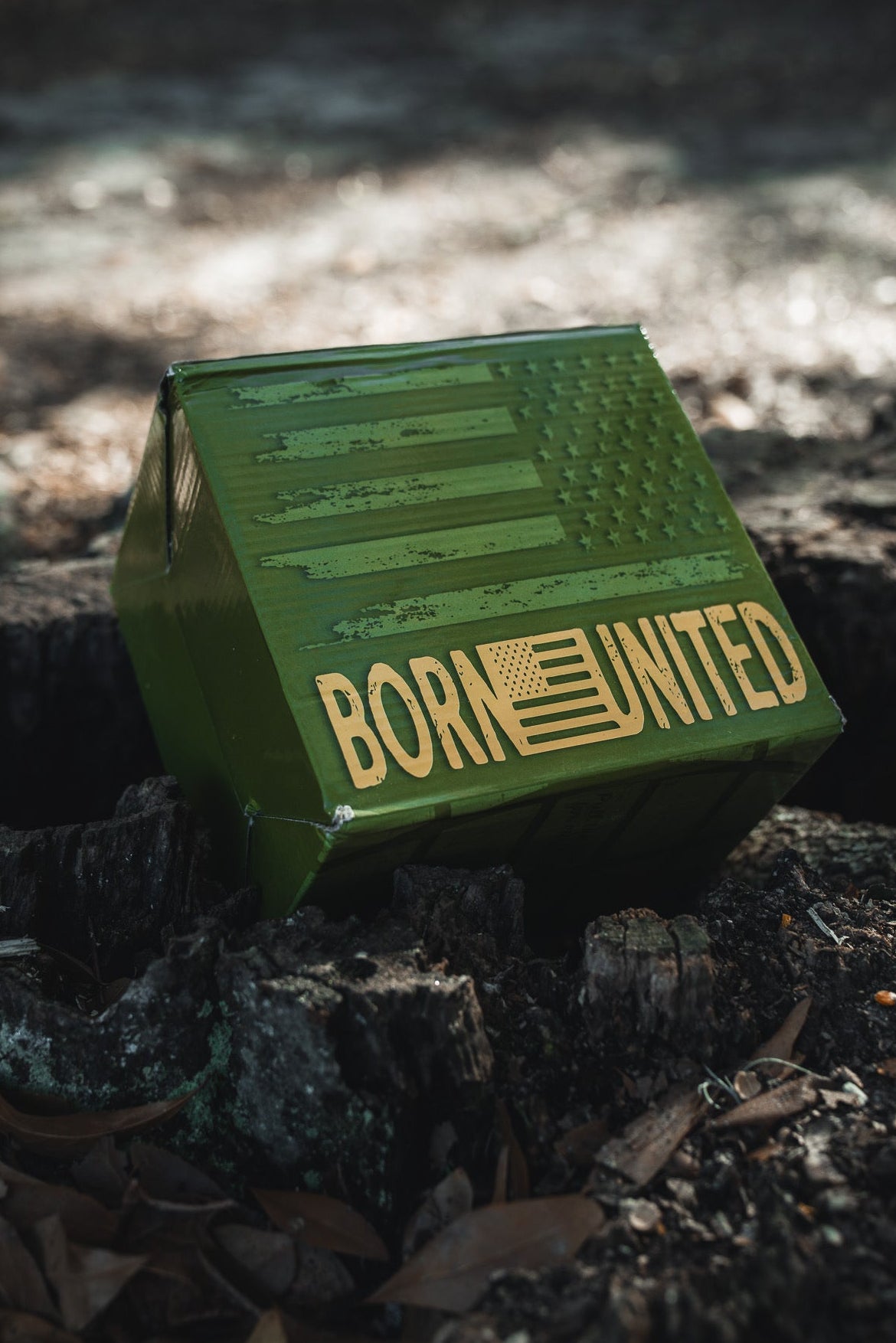 A green box with an American flag and "BORN UNITED" in gold sits on the ground, making it perfect packaging for the BU Sip Sip Boom Mug from Born United Private Label—a bold coffee mug that shines in any outdoor setting.