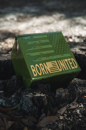 A green box with an American flag and "BORN UNITED" in gold sits on the ground, making it perfect packaging for the BU Sip Sip Boom Mug from Born United Private Label—a bold coffee mug that shines in any outdoor setting.