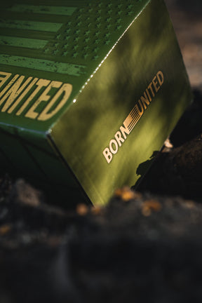 A close-up of the green metallic BU Sip Sip Boom Mug by Born United Private Label, featuring embossed stars and stripes with "BORN UNITED" printed on it, partially lit by sunlight and surrounded by blurred natural scenery.