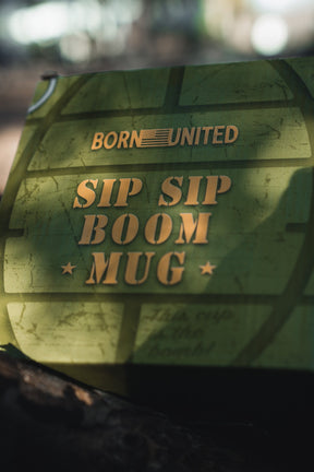 A green BU Sip Sip Boom Mug from Born United Private Label, featuring bold gold “BORN UNITED SIP SIP BOOM MUG” text and a subtle circular design, glows partly in sunlight.