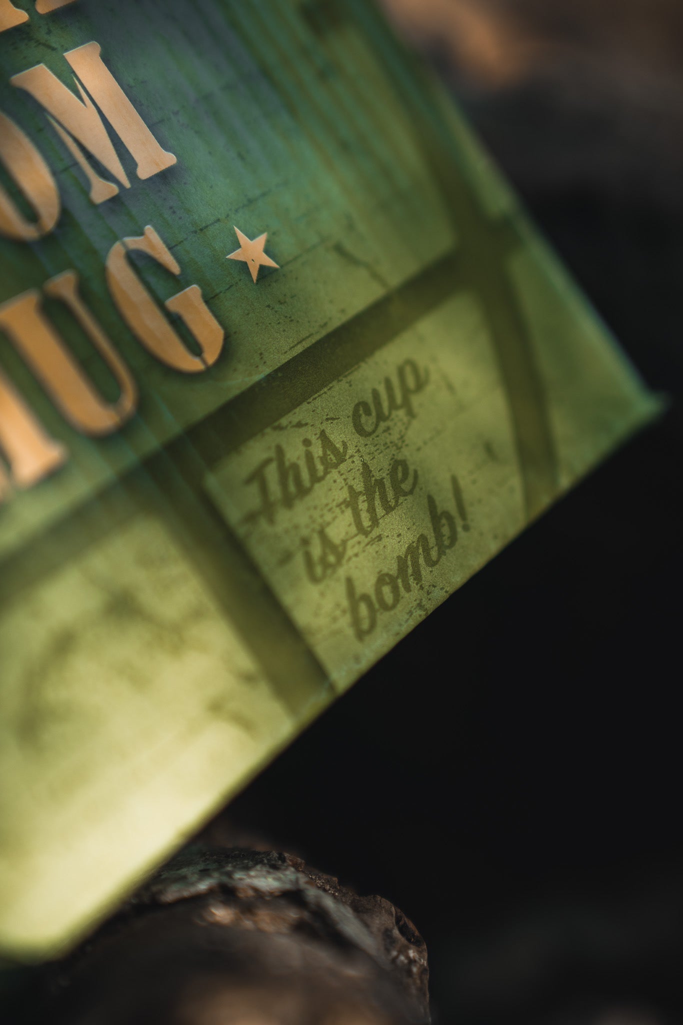 A close-up of the BU Sip Sip Boom Mug from Born United Private Label shows beige text and "This cup is the bomb!" in a playful font, with warm lighting and shadows giving this hand-crafted coffee mug a bold, rustic vibe.