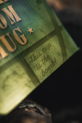 A close-up of the BU Sip Sip Boom Mug from Born United Private Label shows beige text and "This cup is the bomb!" in a playful font, with warm lighting and shadows giving this hand-crafted coffee mug a bold, rustic vibe.
