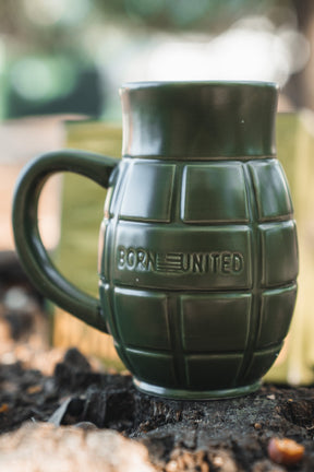 The BU Sip Sip Boom Mug by Born United Private Label, shaped like a green hand grenade with "BORN UNITED" embossed on the side, rests on an outdoor surface against a blurred background.