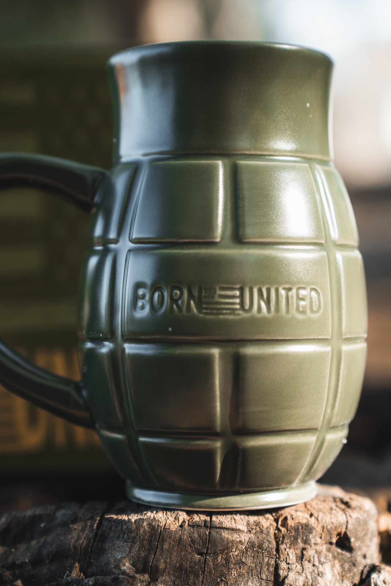The BU Sip Sip Boom Mug by Born United Private Label, an olive green hand grenade-shaped mug with "BORN UNITED" and a flag embossed on its side, sits boldly on a wooden surface.