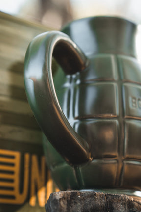 The BU Sip Sip Boom Mug by Born United Private Label features a dark green, hand grenade-shaped design with a handle like a safety lever—set against a blurred gold and green background, it’s an unforgettable conversation starter.