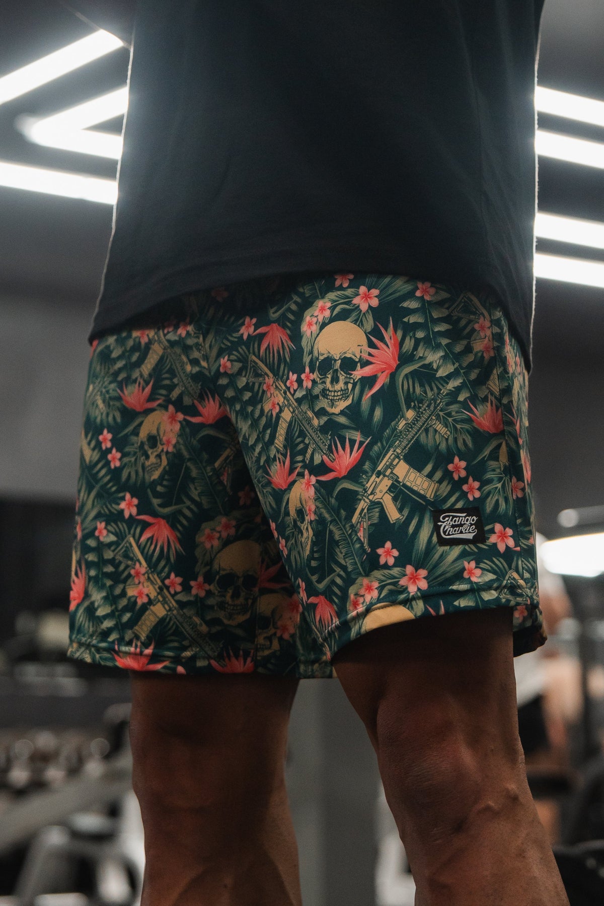 In a well-lit gym filled with modern equipment, a person stands in Mens Performance Training Shorts | 5.5- Aloha AR from Tango Charlie Apparel. The tropical pattern of skulls, leaves, and flowers on the shorts complements their black shirt, creating an ideal setting for performance training.