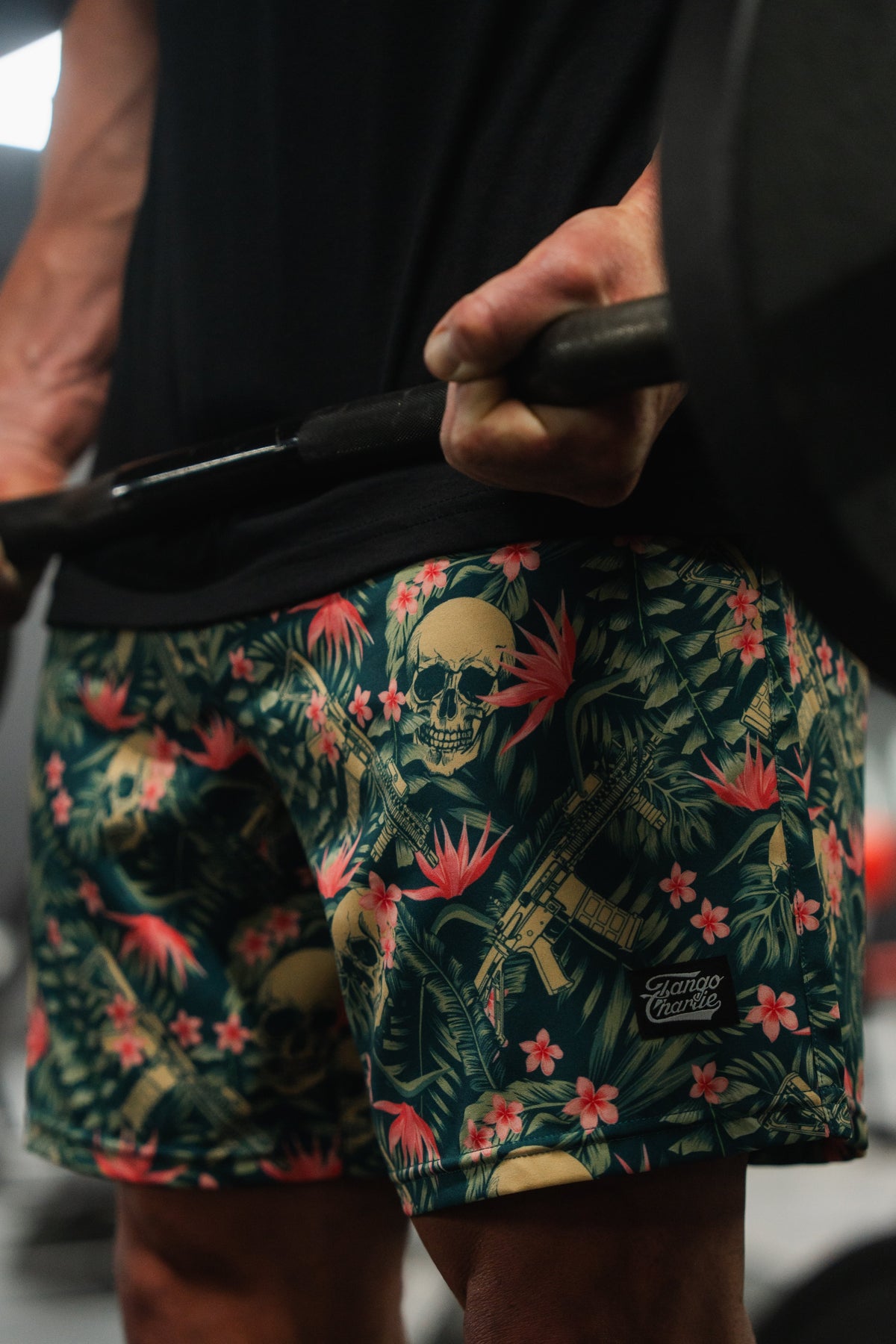 In the gym, someone lifts a barbell, showcasing their Tango Charlie Apparel Mens Performance Training Shorts | 5.5- Aloha AR adorned with vibrant skulls, rifles, and tropical flowers against a blurred background.