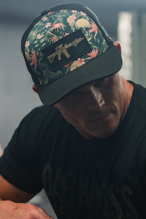 The person wears a sleek black t-shirt and a Tango Charlie Apparel Aloha AR Hydro Snapback Hat with a tropical design, featuring a rifle patch. The blurred background highlights their cool style as shadows partly obscure their face.