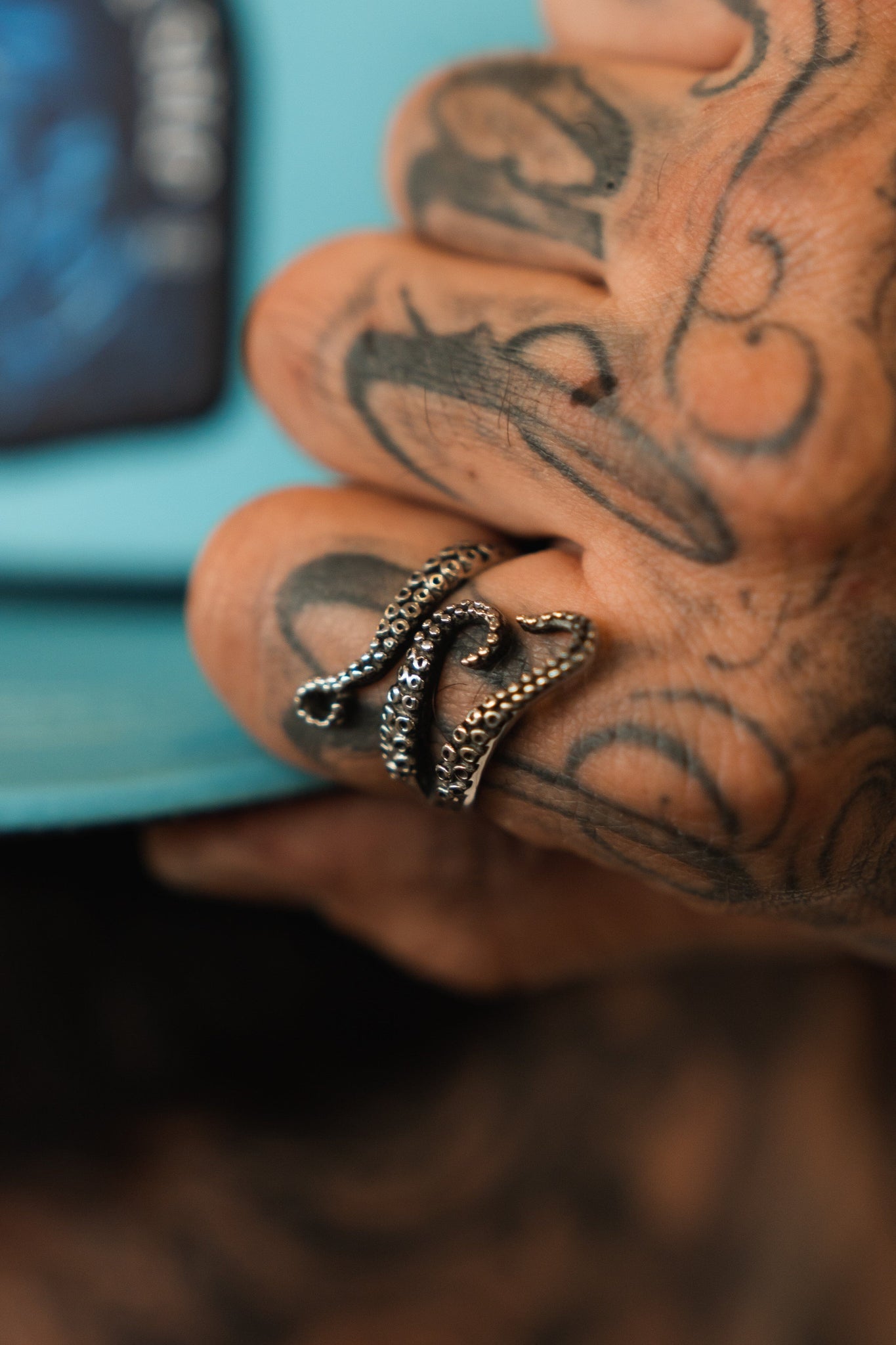 A tattooed hand holds a book, wearing Relentless Betrayals The Kraken Stainless Steel Ring shaped like an octopus tentacle. Its intricate sucker details contrast strikingly with the books blue cover hinting at kraken tales.