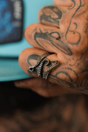 A tattooed hand holds a book, wearing Relentless Betrayals The Kraken Stainless Steel Ring shaped like an octopus tentacle. Its intricate sucker details contrast strikingly with the books blue cover hinting at kraken tales.