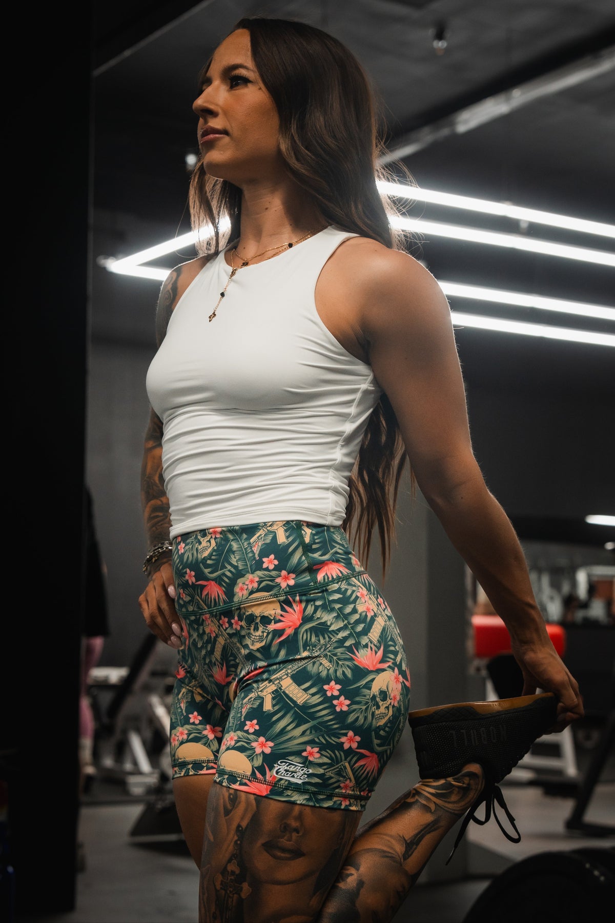 A woman in a white tank top and Tango Charlie Apparels Womens High-Waisted Seamless Front Compression Training Shorts stretches her leg in a dimly lit gym, creating an inspiring workout atmosphere amid the gym equipment.