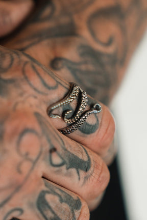 A hand with intricate tattoos wears The Kraken Stainless Steel Ring by Relentless Betrayal, designed like a krakens tentacle. The swirling tattoo patterns enhance the images artistic and nautical theme.