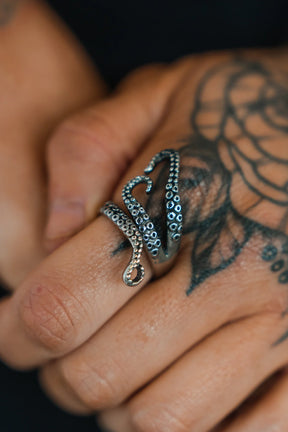 A hand with a detailed flower tattoo on the fingers is adorned with The Kraken Stainless Steel Ring by Relentless Betrayal, a meticulously designed hand-forged piece resembling a krakens tentacle with intricately textured suction cups.