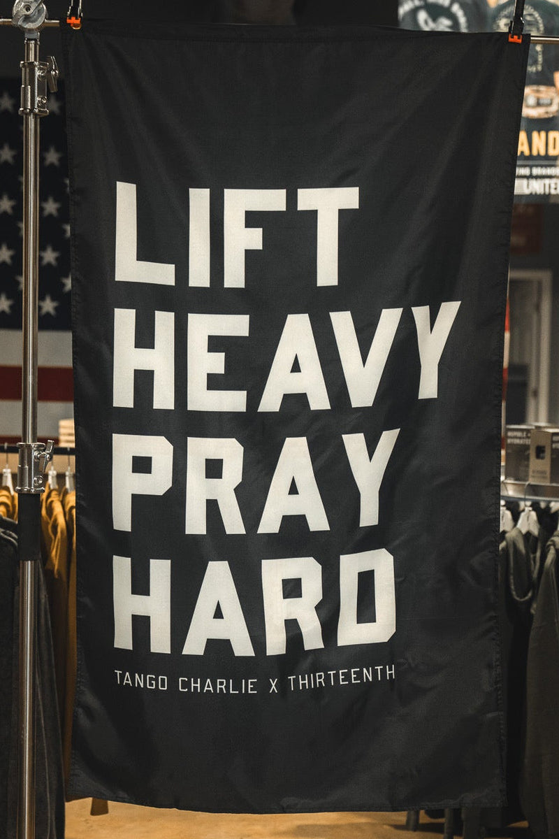 The Lift heavy. Pray hard. - Black Flag by Tango Charlie Apparel hangs in a store, its bold white text and black design standing out among clothing racks and American flags, creating a motivating atmosphere.