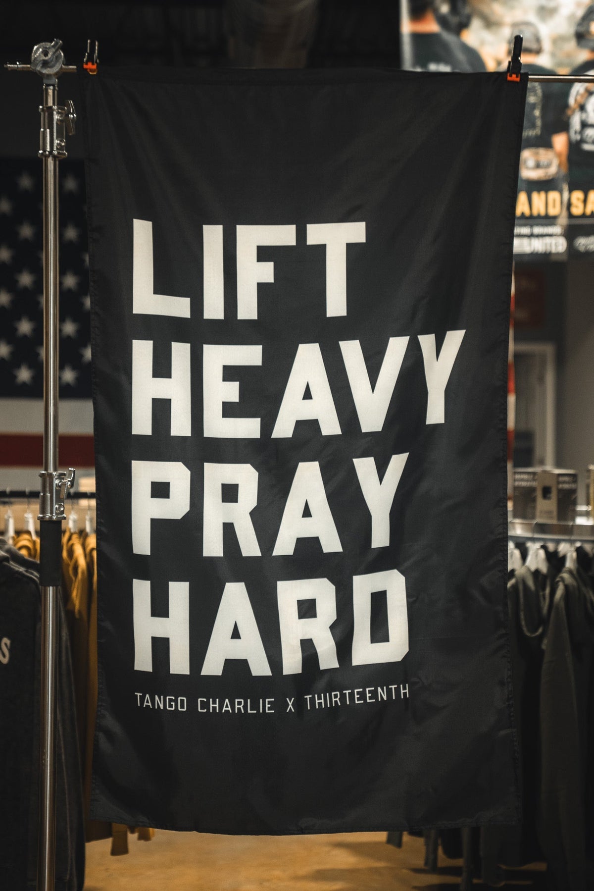The Lift heavy. Pray hard. - Black Flag by Tango Charlie Apparel features bold white text on black polyester and is displayed indoors with clothing racks and an American flag in the background.