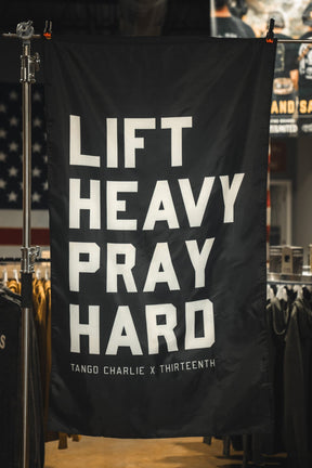 The Lift heavy. Pray hard. - Black Flag by Tango Charlie Apparel features bold white text on black polyester and is displayed indoors with clothing racks and an American flag in the background.