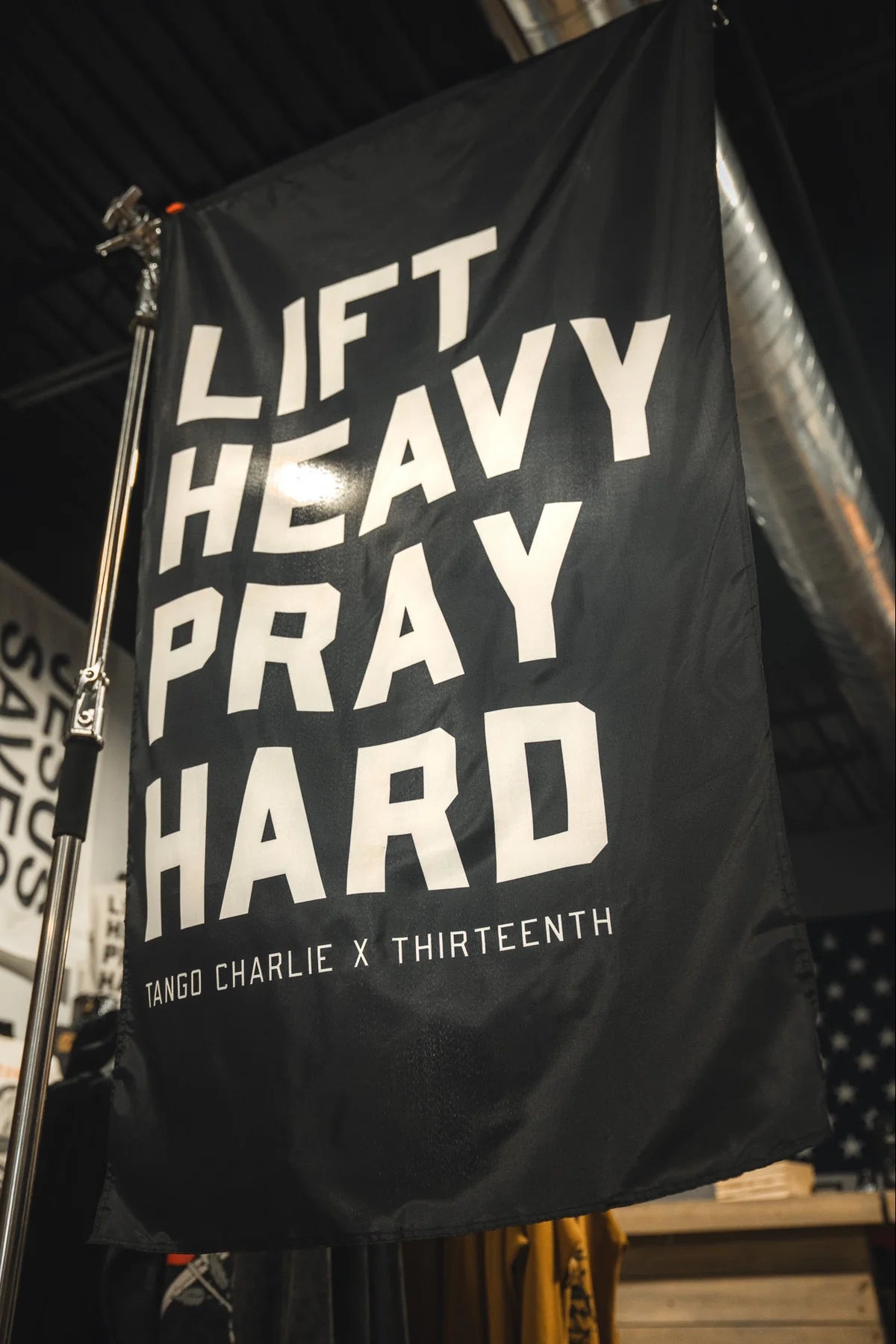 A bold polyester "Lift heavy. Pray hard." black flag from Tango Charlie Apparel hangs as a gym banner, featuring white text beneath exposed industrial pipes. Below, it displays the brand name.