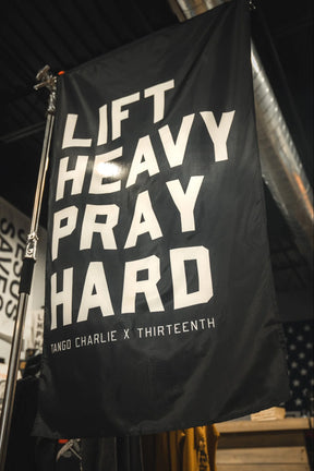 A bold polyester "Lift heavy. Pray hard." black flag from Tango Charlie Apparel hangs as a gym banner, featuring white text beneath exposed industrial pipes. Below, it displays the brand name.