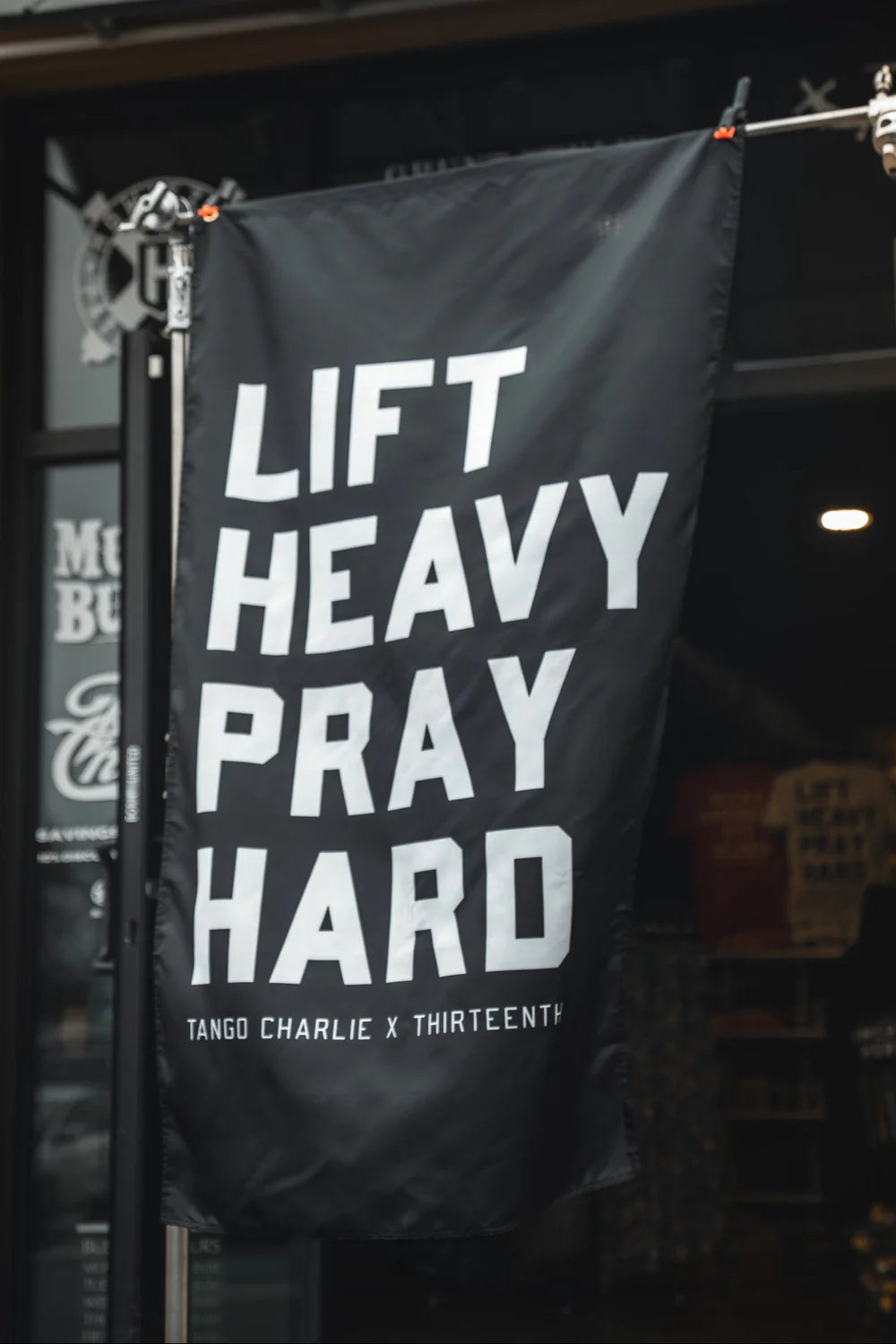 A black Lift heavy. Pray hard. - Black Flag by Tango Charlie Apparel hangs outside a storefront, featuring bold white text and serving as an eye-catching gym banner.