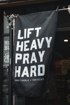 A black Lift heavy. Pray hard. - Black Flag by Tango Charlie Apparel hangs outside a storefront, featuring bold white text and serving as an eye-catching gym banner.