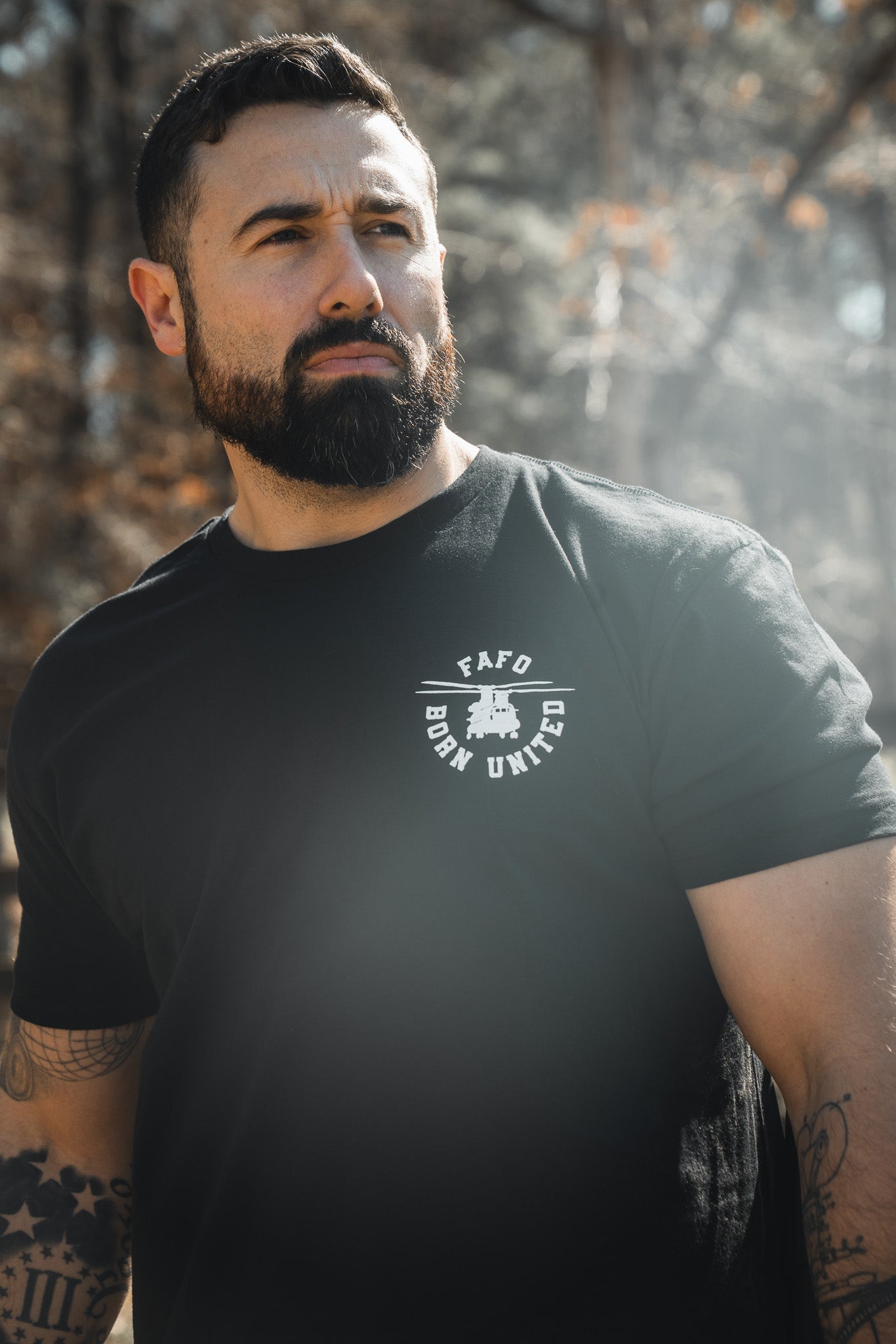 A bearded, tattooed man in a black FAFO - Maduro t-shirt from the Born United Private Label Limited Design collection stands outdoors in sunlight, gazing to the side with a serious look. Blurred trees and a wooden fence are behind him.