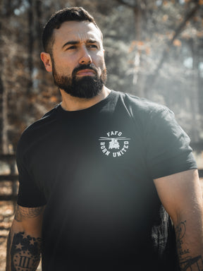 A bearded, tattooed man in a black FAFO - Maduro t-shirt from the Born United Private Label Limited Design collection stands outdoors in sunlight, gazing to the side with a serious look. Blurred trees and a wooden fence are behind him.
