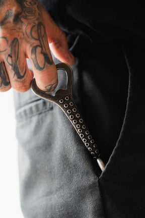A tattooed hand holds the Kraken Bottle Opener by Relentless Betrayal, featuring circular designs on its stainless steel handle, and places it into a black pocket.